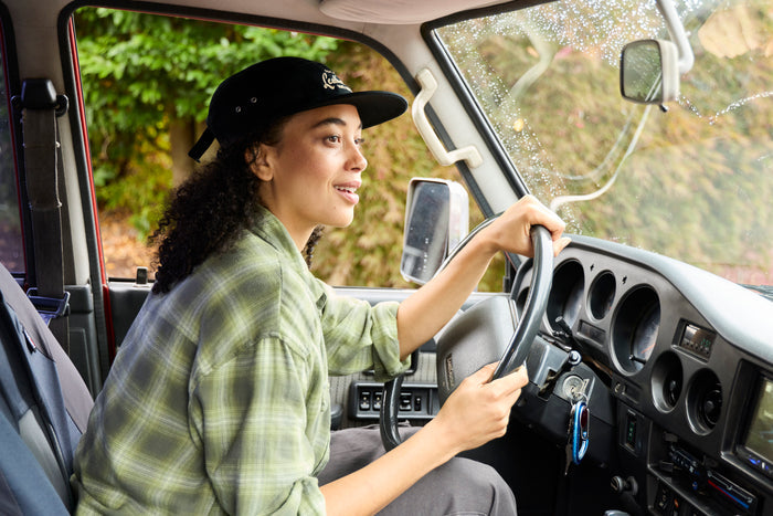 A woman wearing a black Leatherman Vintage 5-Panel Hat driving a vehicle with morning dew on the windshield, smiling.