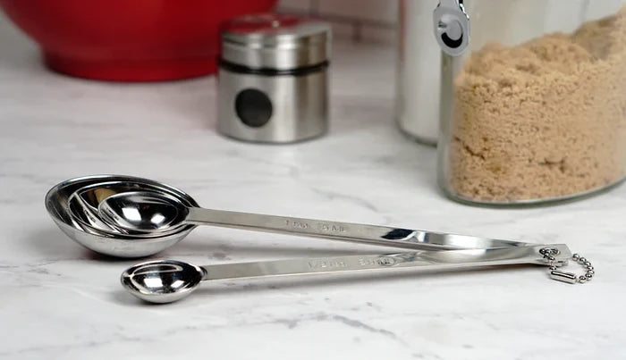 Set of stainless steel measuring spoons on a marble surface with kitchen items in the background.