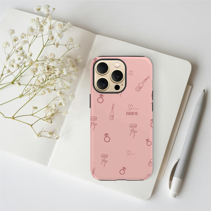 Blush pink personalised bridal tough phone case on a notebook with a pen and white baby’s breath flowers nearby.