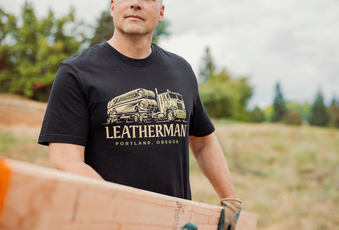 A person wearing a black Leatherman Timber Tee carrying a wooden board outdoors.