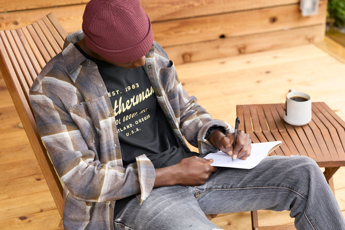A person wearing a black Leatherman Vintage Tee sitting on a wooden chair outdoors, writing in a notebook with a mug of coffee on a nearby table.