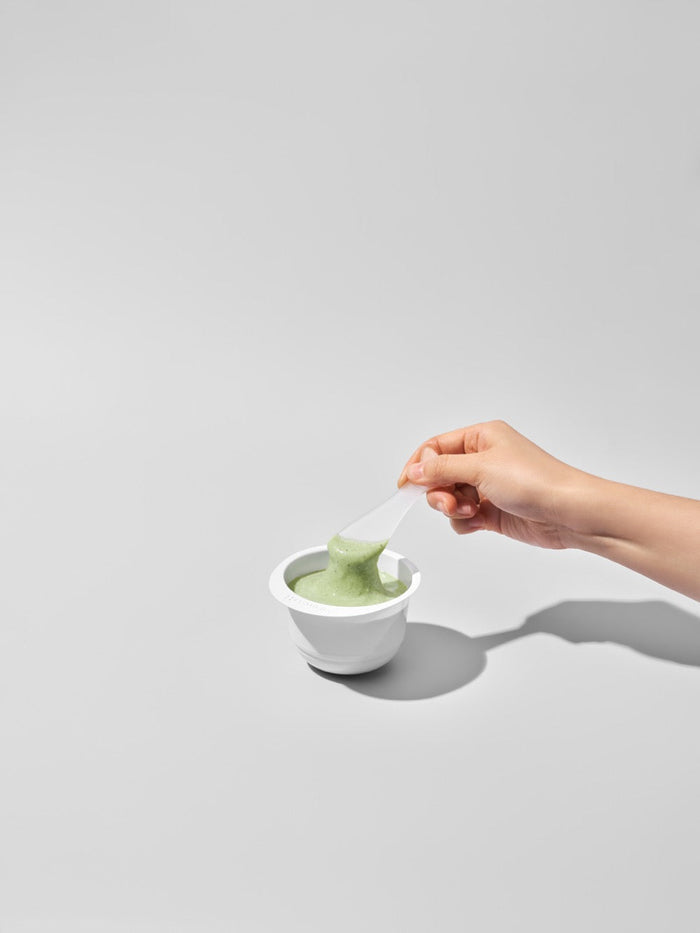 Hand holding a spatula scooping green skincare product from a white bowl, showcasing 3X Boosting Modeling Mask - TeaTree | Leaders for skincare and wellness.