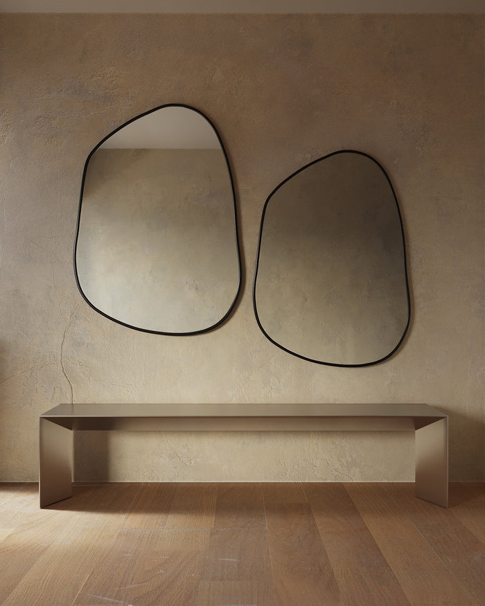 Minimalist interior featuring two asymmetrical wall mirrors with thin black frames mounted above a sleek console table, set against a textured beige wall and warm wood flooring.