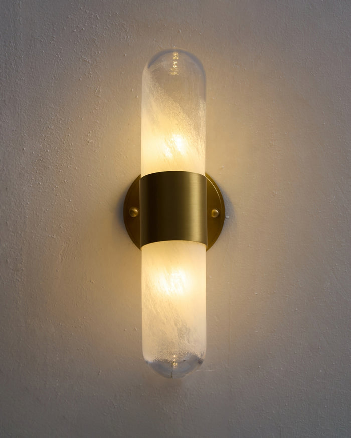 Lit vertical brass wall sconce with a frosted, textured glass tube mounted on a round brass backplate, emitting a warm ambient glow against a textured light-colored wall.