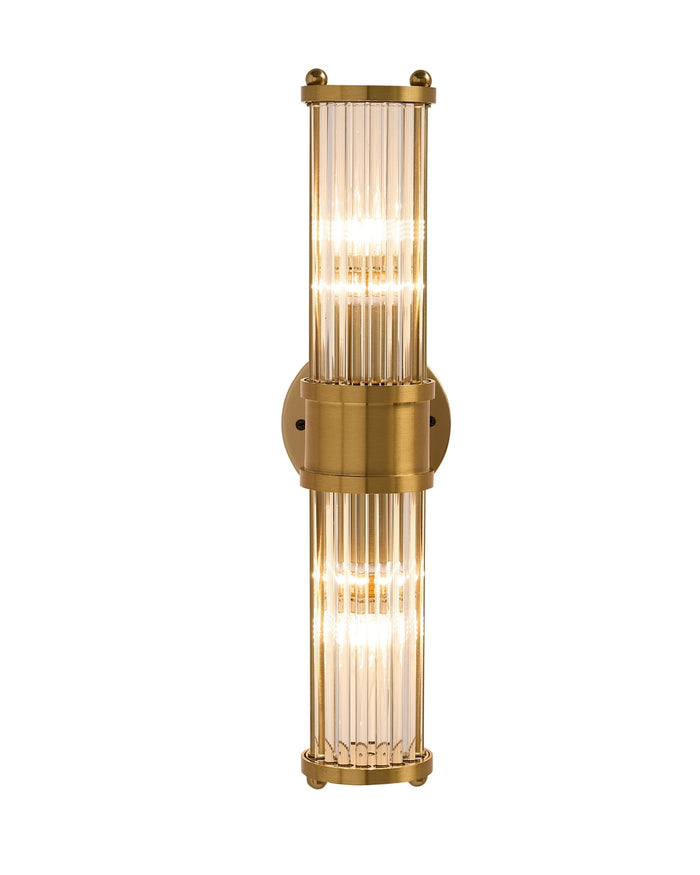 Brimsley II wall sconce with a vertical brass frame and ripple-textured clear glass, featuring two illuminated sections that emit a warm, elegant glow.