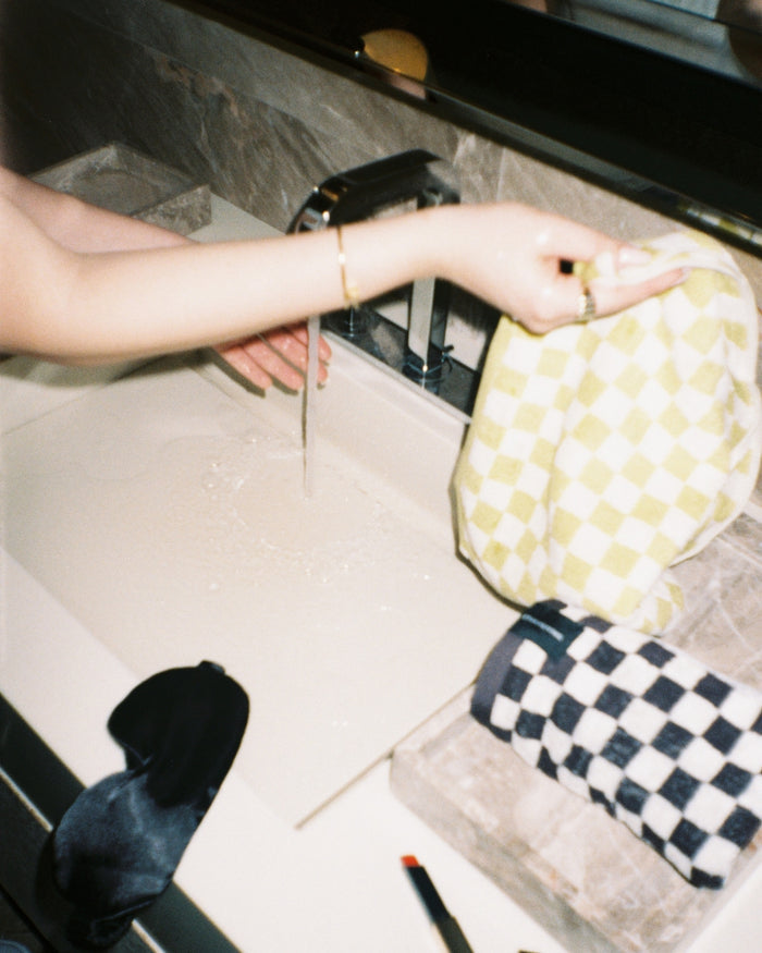 Person holding a yellow checkered towel over a bathroom sink with running water, beside folded checkered towels and a silk sleep mask.