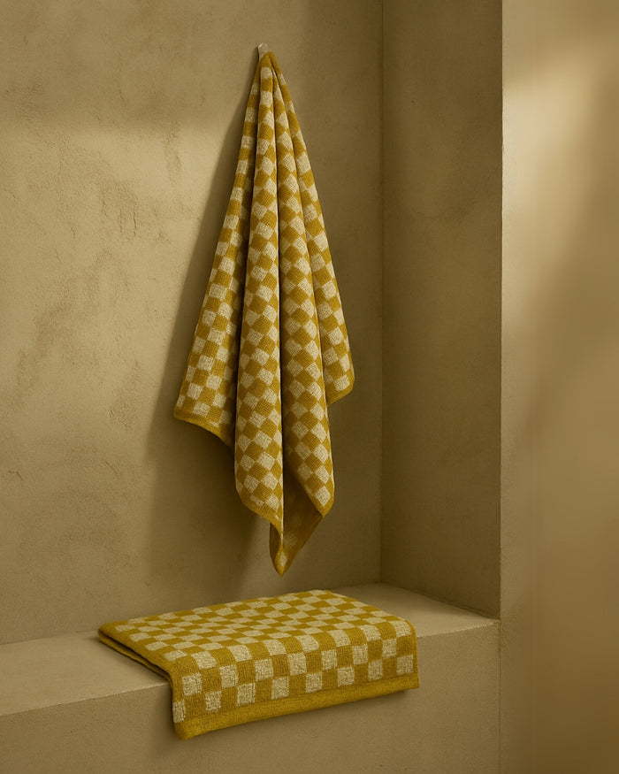 Mustard and white checkered towel hanging on a wall hook, with a folded matching towel placed on a built-in shelf below, photographed in a minimal, neutral-toned interior.