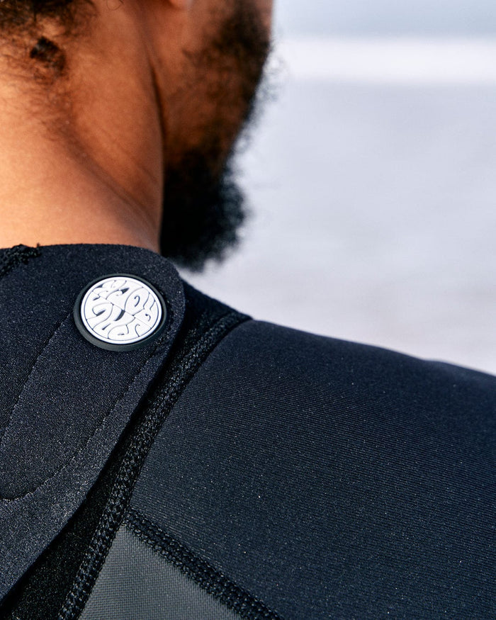 Close-up of a person's neck and shoulder wearing a Saltrock Core - Mens 3/2 Shortie Wetsuit - Black/Blue, featuring a circular emblem with a stylized wave design on the back collar.