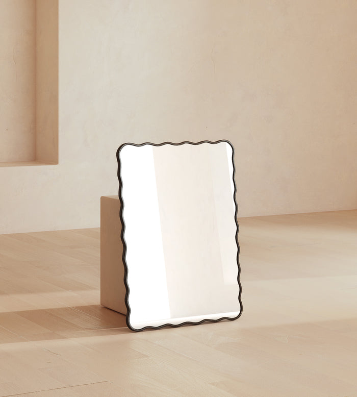 Rectangular wall mirror with a black frame and softly wavy edges, leaning against a wall in a neutral interior setting.