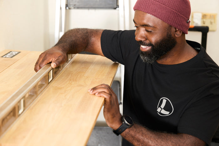 A person wearing a black Leatherman Classic Tee using a level tool to level a wooden shelf indoors.
