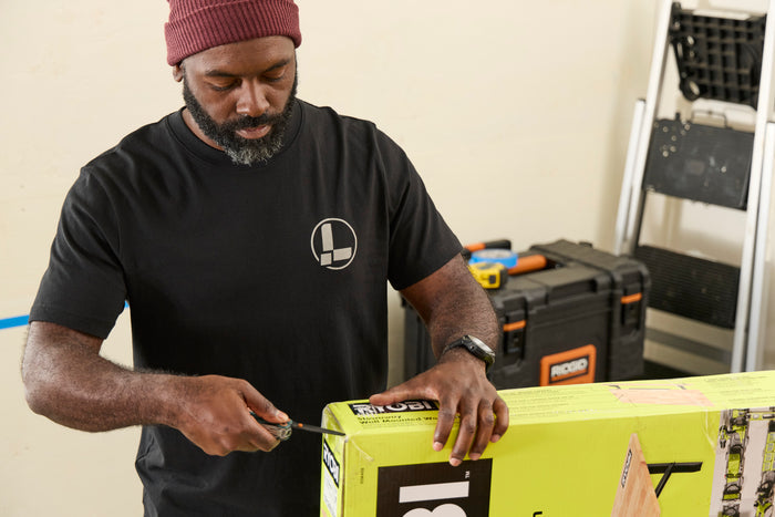 A person wearing a black Leatherman Classic Tee using the blade on a Wave Alpha in Cascadia color to open a large box in a workshop while standing near a ladder and tool case.