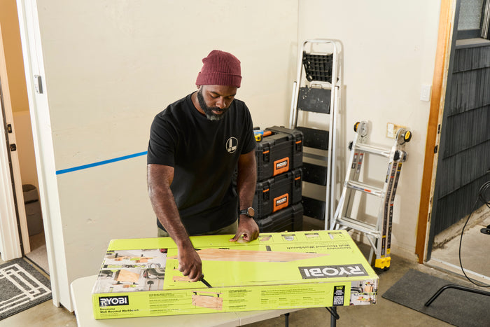 A person wearing a black Leatherman Classic Tee using the blade on a Leatherman multi-tool to open a large box that is laying on top of a folding table.