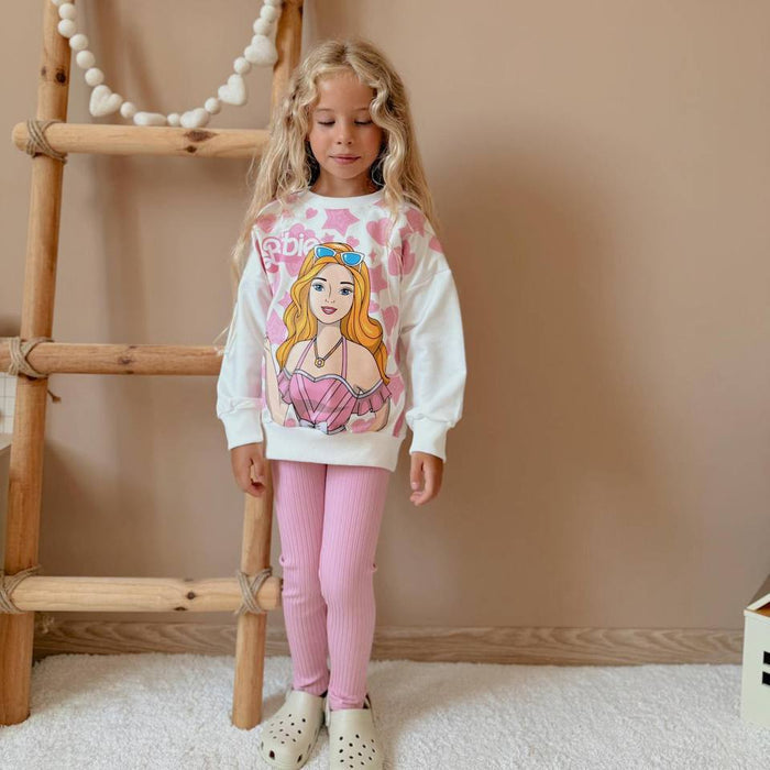 Girl wearing a Barbie graphic sweatshirt with pink leggings, standing indoors beside a wooden ladder on a neutral background.