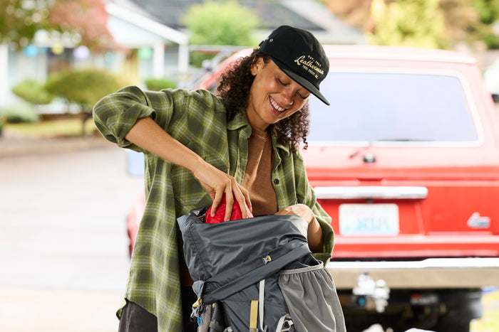 A woman wearing a black Leatherman Vintage 5 Panel Hat packing a gray hiking backpack near a red vehicle.