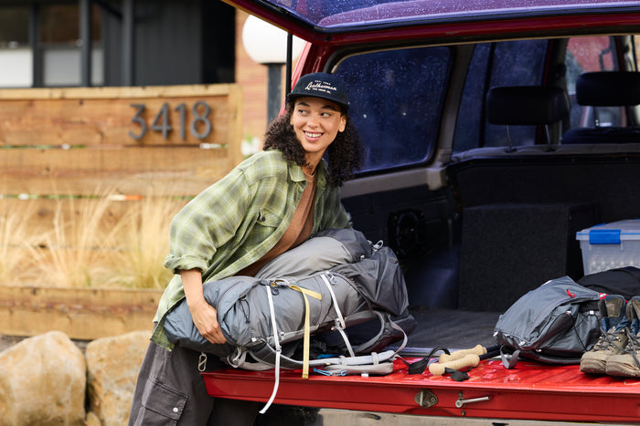 A woman wearing a black Leatherman Vintage 5-Panel Hat loading camping gear into the back of a red vehicle, smiling while preparing for an outdoor trip.