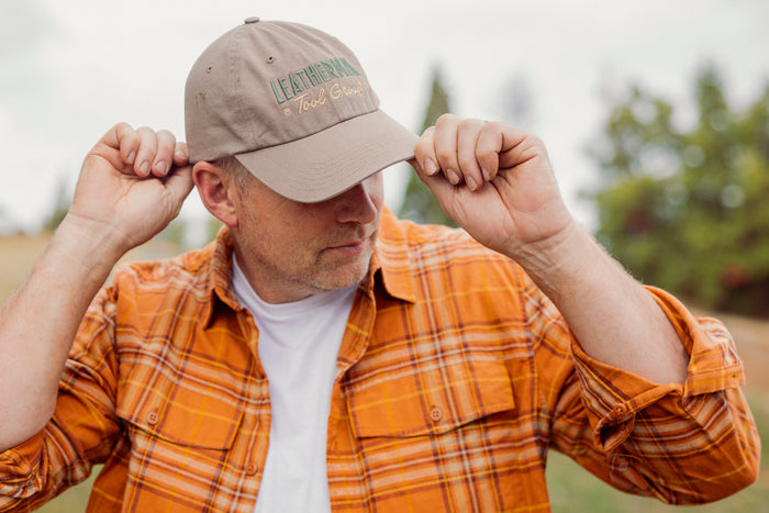 A person wearing a Leatherman Dad Hat in Driftwood color adjusting the brim while outdoors, dressed in an orange plaid shirt.