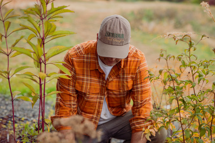 A person wearing a driftwood colored Leatherman Dad Hat tending to plants outdoors while dressed in an orange plaid shirt.