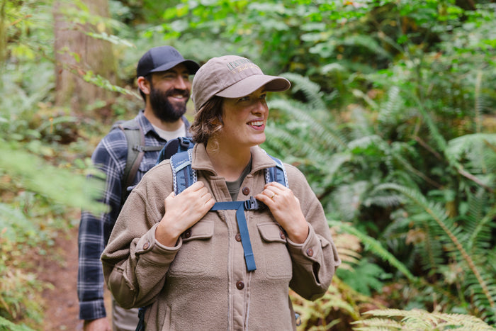 A woman wearing a Leatherman Dad Hat in Driftwood color hiking through a forest with a backpack smiling as another hiker follows behind.