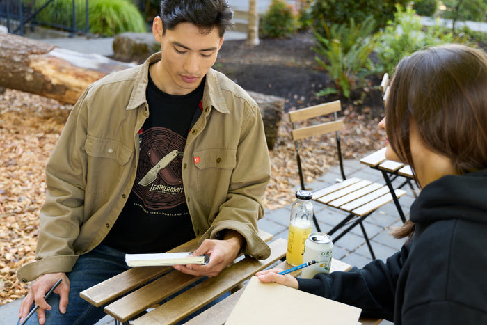 A man sitting at a table wearing a black Leatherman Blade & Grain tee with a woman wearing a black hoodie, both equipped with a notebook and pencil.