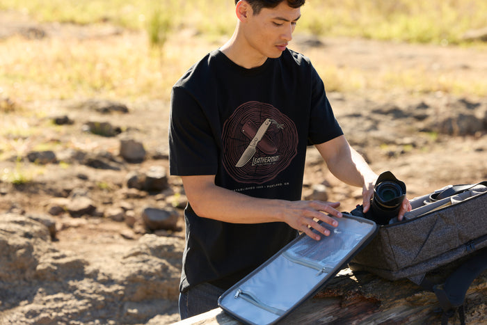 A person wearing a black Leatherman Blade & Grain tee pulling a camera out of a camera pack.