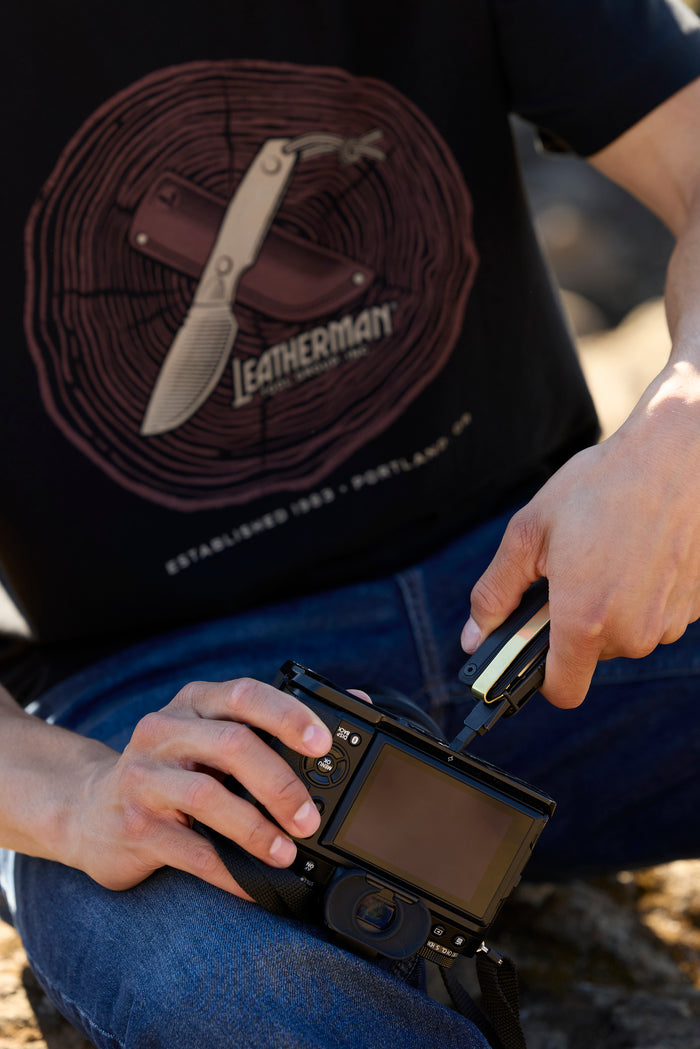 A person using a Leatherman Wave Alpha bit driver to tighten a bolt on a camera while wearing a black Blade & Grain Tee.