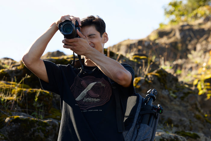 A photographer wearing a black Leatherman Blade & Grain Tee while taking a pictures outdoors.