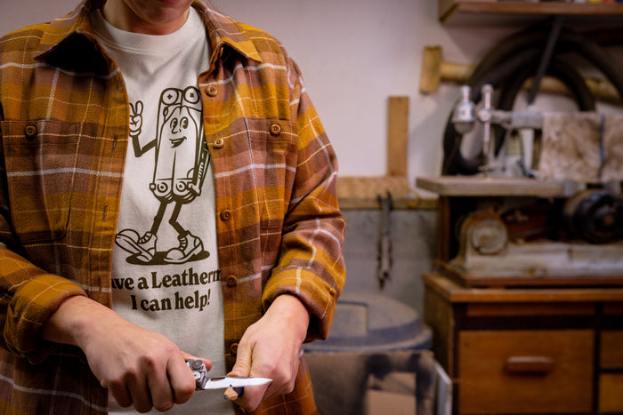 A person wearing a natural-colored Leatherman Mr. Wave Tee with a cartoon multitool design using a Leatherman multitool to sharpen a pencil.
