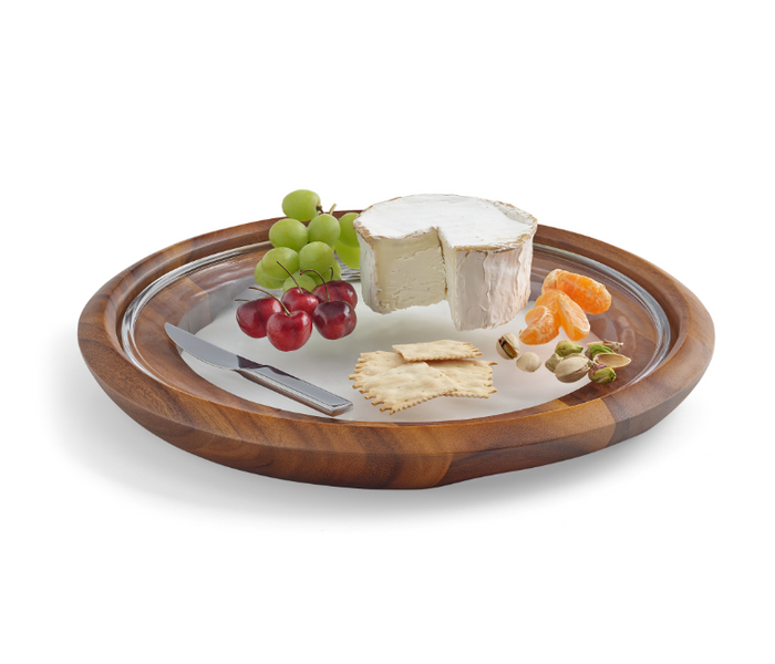 An acacia wood cheese tray with a glass server and stainless steel knife, accompanied by grapes and pieces of cheese.