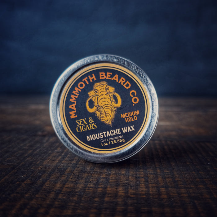 Mammoth Beard Co. Sex & Cigars Moustache Wax tin in focus on a rustic surface, showcasing the bold gold mammoth logo and “Medium Hold” marking on the label.