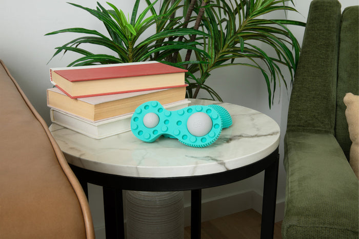 Neuro Roller | RAD wellness tool in turquoise with textured surface resting on a white marble round side table beside stacked books and green plants in a cozy living room setting.
