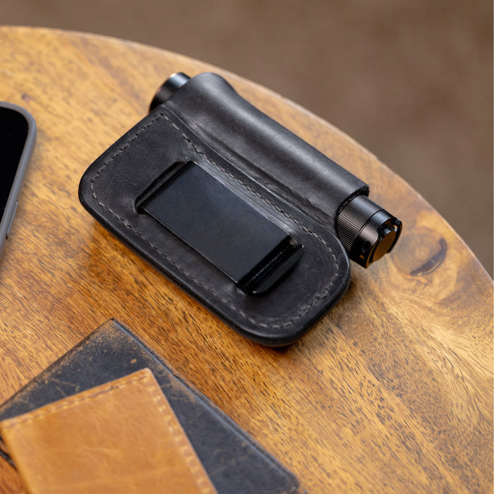 Black leather sheath with belt clip holding a small cylindrical flashlight on a wooden surface