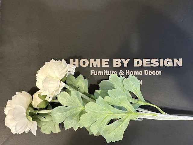 An artificial ranunculus spray with cream-colored flowers, placed on a surface with a black background featuring the text 'HOME BY DESIGN Furniture & Home Decor'.