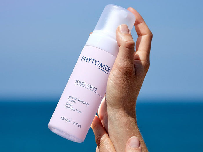 Hand holding a bottle of Rosée Visage Gentle Cleansing Foam | Phytomer skincare product against a clear blue sky and sea background, emphasizing gentle cleansing and freshness.