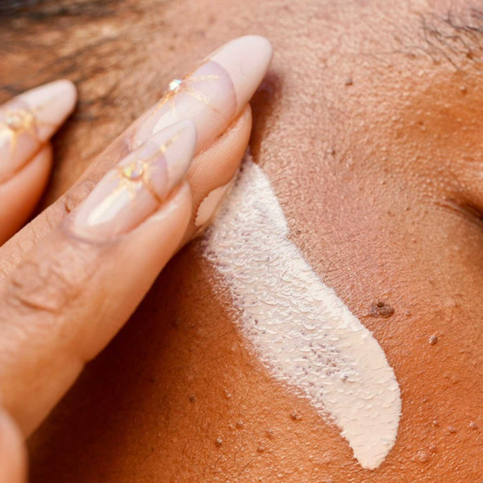 Close-up of a person applying a strip of SkinSuit Daily Protector SPF 50 | Skin Authority sunscreen on their cheek, showcasing smooth skin and detailed nail art.