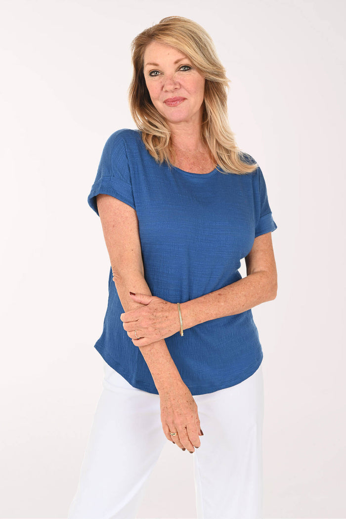 A woman modeling a short-sleeve blue top with white pants against a plain white studio background.
