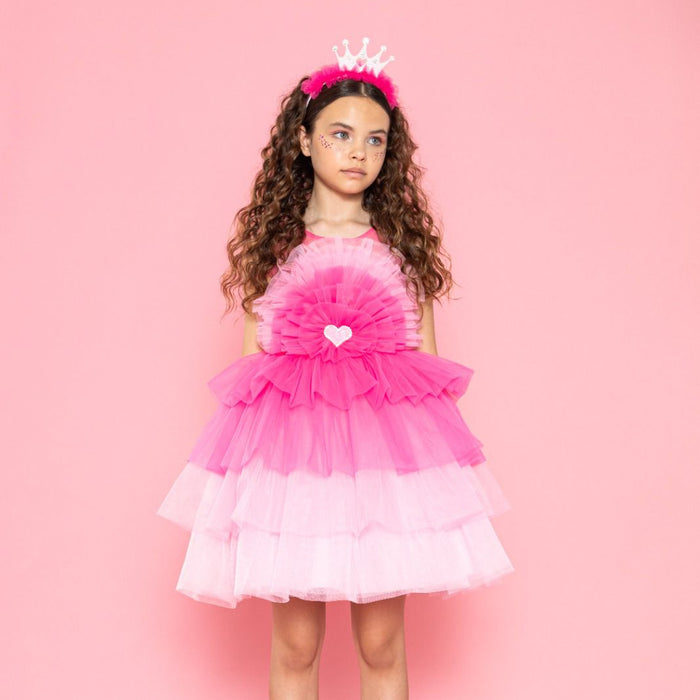 Girl in a layered pink tulle dress with a heart detail, wearing a crown headband, standing against a pink background.