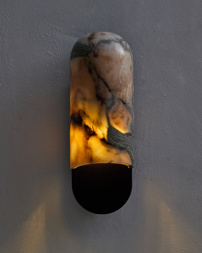 Marble wall sconce mounted on a textured gray wall, featuring a rounded vertical form with natural beige, gray, and amber veining, softly illuminated from within above a gunblack metal base.