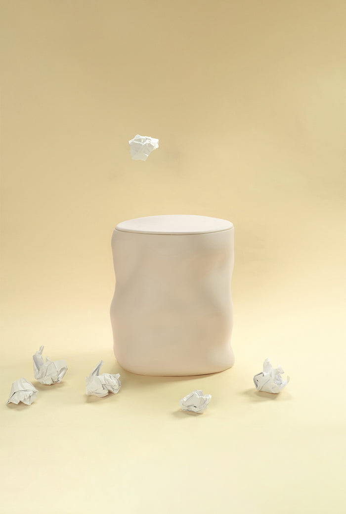 A beige sculptural dustbin placed against a light yellow background, with several crumpled pieces of white paper on the floor around it and one piece suspended above the bin.