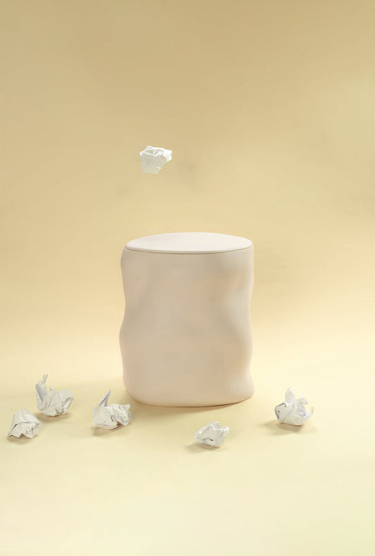 A beige sculptural dustbin placed against a light yellow background, with several crumpled pieces of white paper on the floor around it and one piece suspended above the bin.