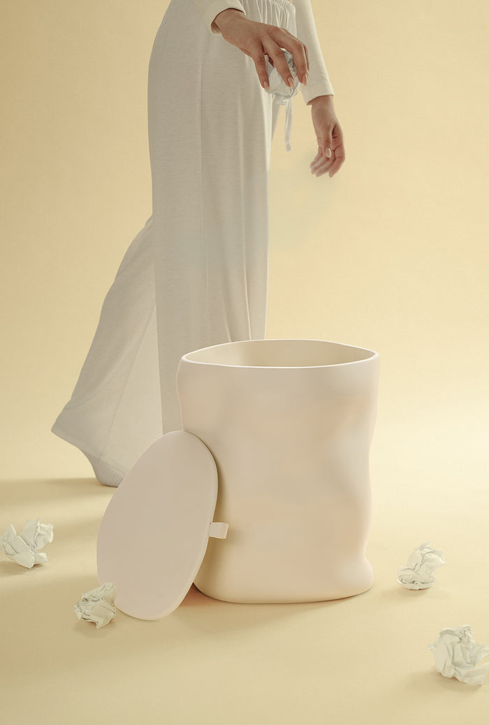 A beige sculptural dustbin shown with its lid placed to the side, as a person drops a crumpled piece of white paper into the open bin, against a light yellow background.