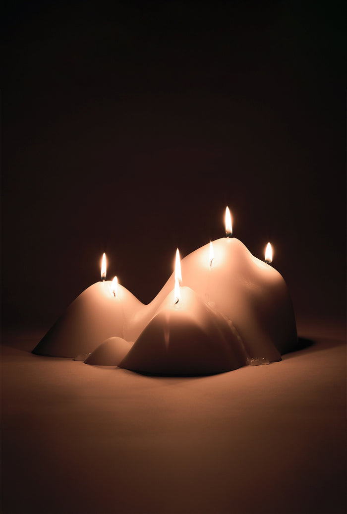 White sculptural candle with an organic, mountain-inspired form and multiple lit wicks, softly illuminated against a dark background.
