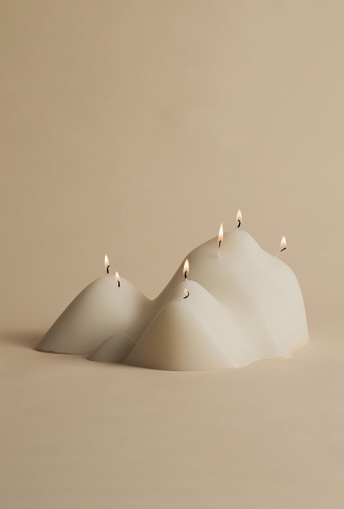 White sculptural candle with an organic, mountain-like form and multiple lit wicks, resting on a neutral surface against a softly lit background.