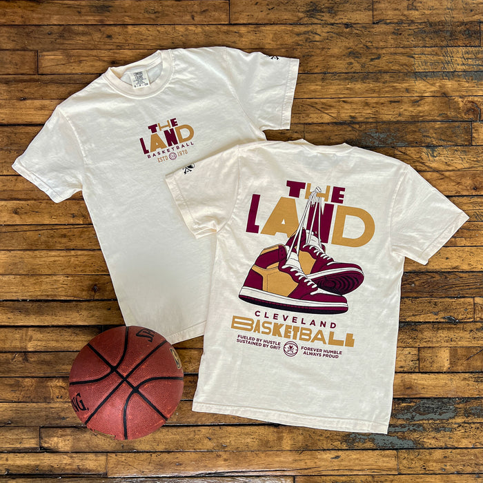 Two white t-shirts with 'The Land' and basketball graphics on a wooden surface, accompanied by a basketball.
