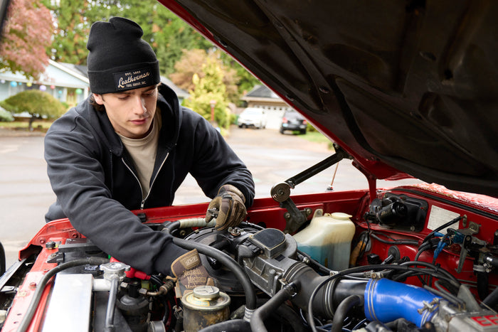 A person wearing a black Leatherman Vintage Beanie working under the hood of a red vehicle in a residential driveway.
