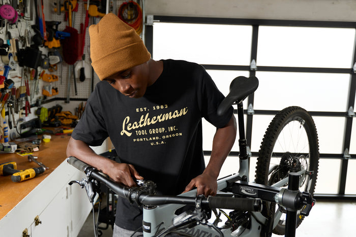 A person wearing a black Leatherman Vintage Tee working on a mountain bike in a garage workshop, surrounded by tools and equipment.