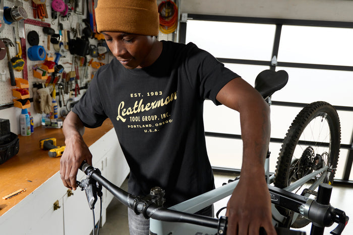 A person wearing a black Leatherman Vintage Tee working on a mountain bike in a garage workshop, surrounded by tools and equipment.