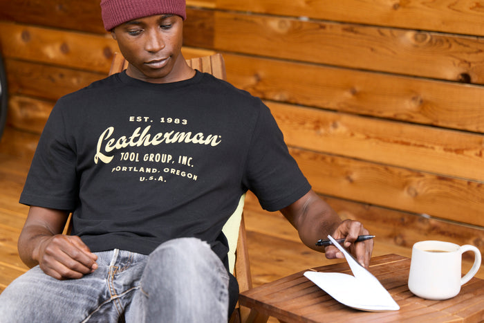 A person wearing a black Leatherman Vintage Tee sitting on a wooden deck chair, opening a notebook beside a mug of coffee.