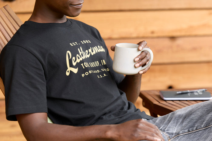 A person wearing a black Leatherman Vintage Tee relaxing on a wooden deck chair, holding a white mug with a notebook and pen on a small table nearby.